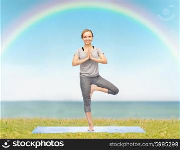 fitness, sport, people and healthy lifestyle concept - woman making yoga in tree pose on mat over blue sky, rainbow and sea background