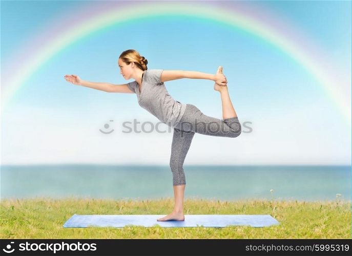fitness, sport, people and healthy lifestyle concept - woman making yoga in lord of the dance pose on mat over blue sky, rainbow and sea background