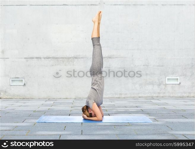 fitness, sport, people and healthy lifestyle concept - woman making yoga in headstand pose on mat over urban street background
