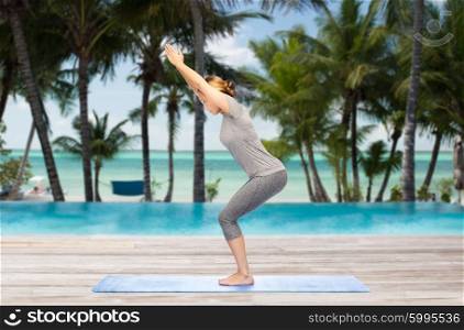 fitness, sport, people and healthy lifestyle concept - woman making yoga in chair pose on mat over hotel resort pool on tropical beach background