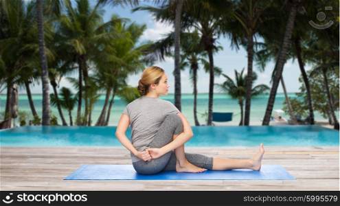 fitness, sport, people and healthy lifestyle concept - woman making yoga in twist pose on mat over hotel resort pool on tropical beach background