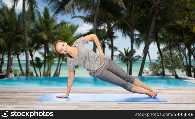 fitness, sport, people and healthy lifestyle concept - woman making yoga in side plank pose on mat over hotel resort pool on tropical beach background