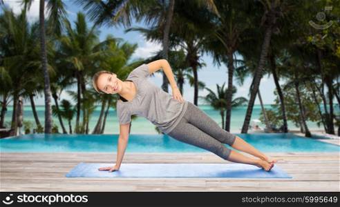 fitness, sport, people and healthy lifestyle concept - woman making yoga in side plank pose on mat over hotel resort pool on tropical beach background