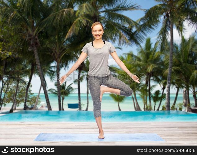 fitness, sport, people and healthy lifestyle concept - woman making yoga in tree pose on mat over hotel resort pool on tropical beach background