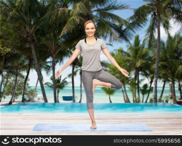 fitness, sport, people and healthy lifestyle concept - woman making yoga in tree pose on mat over hotel resort pool on tropical beach background