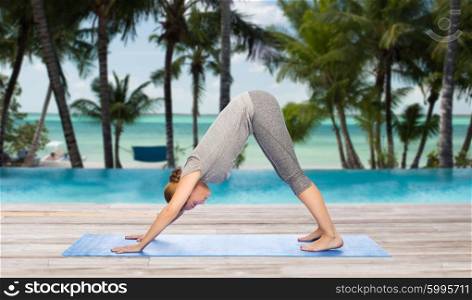 fitness, sport, people and healthy lifestyle concept - woman making yoga in downward facing dog pose on mat over hotel resort pool on tropical beach background