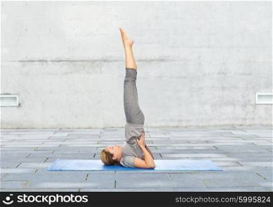 fitness, sport, people and healthy lifestyle concept - woman making yoga in shoulderstand pose on mat over urban street background