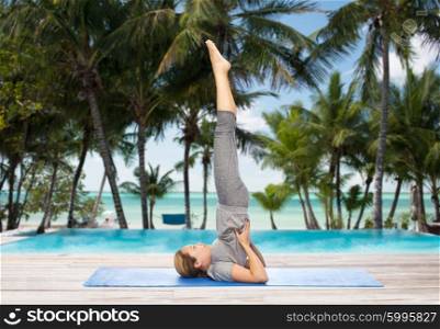 fitness, sport, people and healthy lifestyle concept - woman making yoga in shoulderstand pose on mat over hotel resort pool on tropical beach background