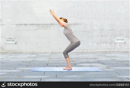 fitness, sport, people and healthy lifestyle concept - woman making yoga in chair pose on mat over urban street background