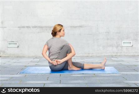 fitness, sport, people and healthy lifestyle concept - woman making yoga in twist pose on mat over urban street background