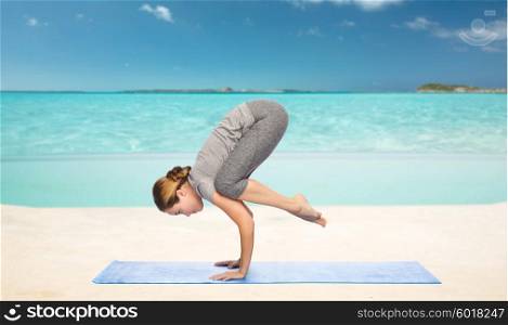 fitness, sport, people and healthy lifestyle concept - woman making yoga in crane pose on mat over sea and sky background