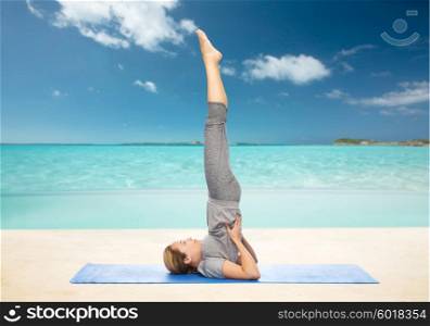 fitness, sport, people and healthy lifestyle concept - woman making yoga in shoulderstand pose on mat over beach background