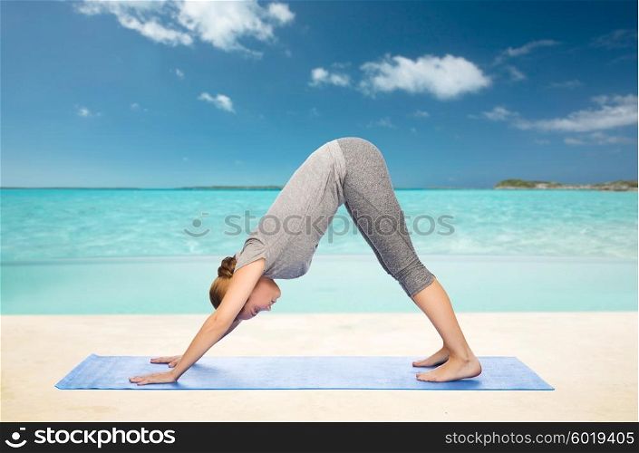 fitness, sport, people and healthy lifestyle concept - woman making yoga in downward facing dog pose on mat over beach background