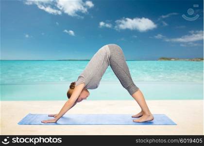 fitness, sport, people and healthy lifestyle concept - woman making yoga in downward facing dog pose on mat over beach background