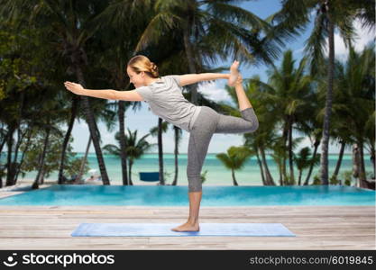 fitness, sport, people and healthy lifestyle concept - woman making yoga in lord of the dance pose on mat over hotel resort pool on tropical beach background