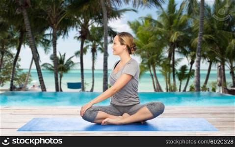 fitness, sport, people and healthy lifestyle concept - woman making yoga in twist pose on mat over hotel resort pool on tropical beach background
