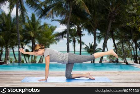 fitness, sport, people and healthy lifestyle concept - woman making yoga in balancing table pose on mat over hotel resort pool on tropical beach background