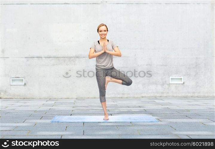 fitness, sport, people and healthy lifestyle concept - woman making yoga in tree pose on mat over urban street background
