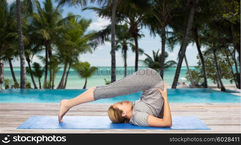 fitness, sport, people and healthy lifestyle concept - woman making yoga in plow pose on mat over hotel resort pool on tropical beach background