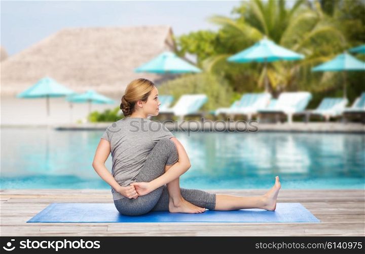 fitness, sport, people and healthy lifestyle concept - woman making yoga in twist pose on mat over beach and swimming pool background