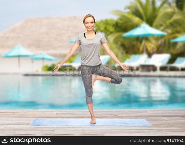 fitness, sport, people and healthy lifestyle concept - woman making yoga in tree pose on mat over beach and swimming pool background