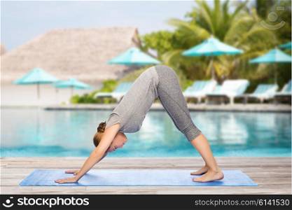 fitness, sport, people and healthy lifestyle concept - woman making yoga in downward facing dog pose on mat over beach and swimming pool background