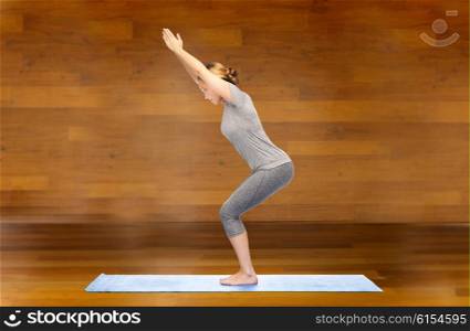 fitness, sport, people and healthy lifestyle concept - woman making yoga in chair pose on mat over wooden room background