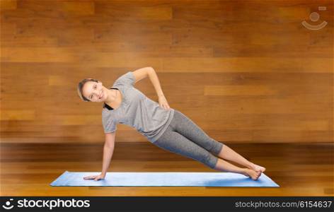fitness, sport, people and healthy lifestyle concept - woman making yoga in side plank pose on mat over wooden room background