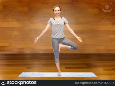 fitness, sport, people and healthy lifestyle concept - woman making yoga in tree pose on mat over wooden room background