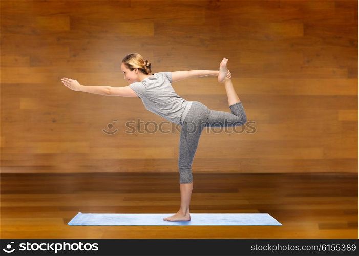 fitness, sport, people and healthy lifestyle concept - woman making yoga in lord of the dance pose on mat over wooden room background