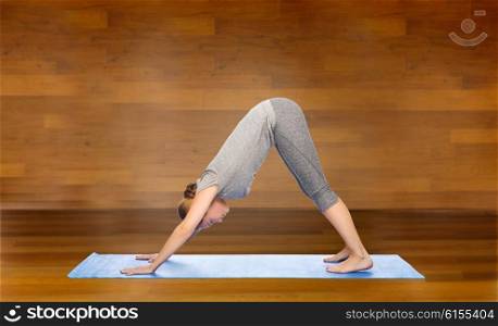 fitness, sport, people and healthy lifestyle concept - woman making yoga in downward facing dog pose on mat over wooden room background