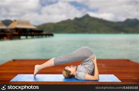 fitness, sport, people and healthy lifestyle concept - woman making yoga in plow pose on wooden pier over island beach and bungalow background. woman making yoga in plow pose on mat outdoors