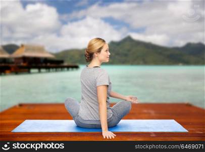fitness, sport, people and healthy lifestyle concept - woman making yoga in twist pose on wooden pier over island beach and bungalow background. woman making yoga in twist pose on mat outdoors
