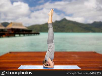 fitness, sport, people and healthy lifestyle concept - woman making yoga in headstand pose on wooden pier over island beach and bungalow background. woman making yoga in headstand pose outdoors