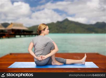 fitness, sport, people and healthy lifestyle concept - woman making yoga in twist pose on wooden pier over island beach and bungalow background. woman making yoga in twist pose on mat outdoors