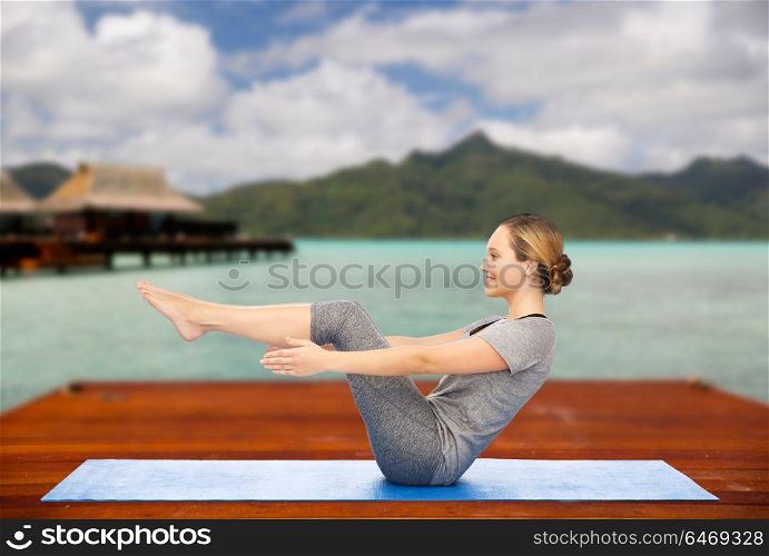 fitness, sport, people and healthy lifestyle concept - woman making yoga in half-boat pose on wooden pier over island beach and bungalow background. woman making yoga half-boat pose on mat outdoors