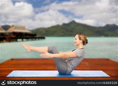 fitness, sport, people and healthy lifestyle concept - woman making yoga in half-boat pose on wooden pier over island beach and bungalow background. woman making yoga half-boat pose on mat outdoors