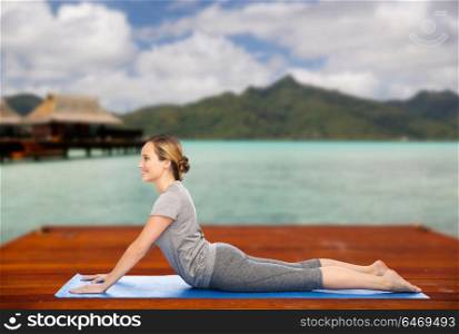 fitness, sport, people and healthy lifestyle concept - woman making yoga in dog pose on wooden pier over island beach and bungalow background. woman making yoga in dog pose on mat outdoors