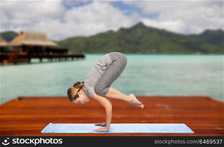 fitness, sport, people and healthy lifestyle concept - woman making yoga in crane pose on wooden pier over island beach and bungalow background. woman making yoga in crane pose on mat outdoors