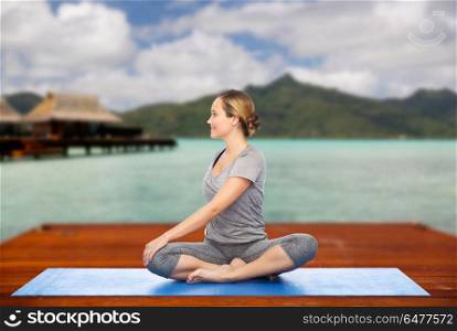 fitness, sport, people and healthy lifestyle concept - woman making yoga in twist pose on wooden pier over island beach and bungalow background. woman making yoga in twist pose on mat outdoors. woman making yoga in twist pose on mat outdoors