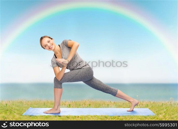 fitness, sport, people and healthy lifestyle concept - woman making yoga low angle lunge pose on mat over blue sky, rainbow and sea background