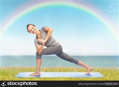 fitness, sport, people and healthy lifestyle concept - woman making yoga low angle lunge pose on mat over blue sky, rainbow and sea background