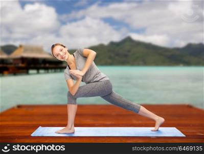 fitness, sport, people and healthy lifestyle concept - woman making yoga low angle lunge pose on wooden pier over island beach and bungalow background. woman making yoga low angle lunge pose outdoors