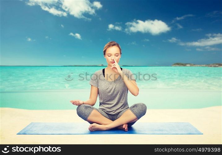 fitness, sport, people and healthy lifestyle concept - woman making yoga meditation in lotus pose on mat over sea and sky background. woman meditating in lotus yoga pose on beach