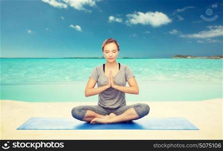 fitness, sport, people and healthy lifestyle concept - woman making yoga meditation in lotus pose on mat over sea and sky background. woman meditating in lotus yoga pose on beach