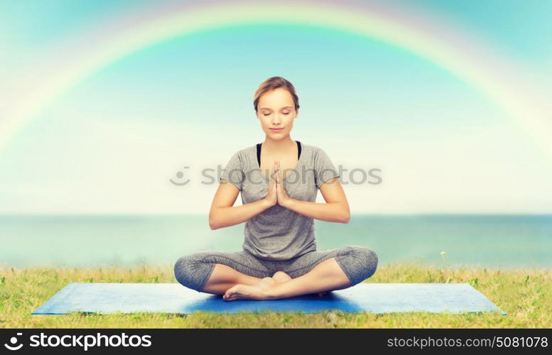 fitness, sport, people and healthy lifestyle concept - woman making yoga meditation in lotus pose on mat over blue sky, rainbow and sea background. woman making yoga meditation in lotus pose on mat