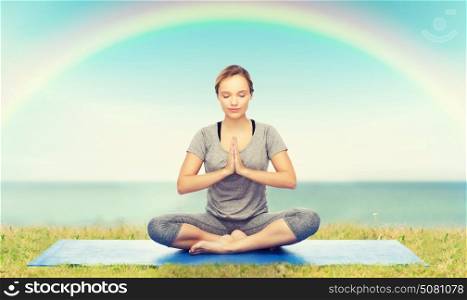 fitness, sport, people and healthy lifestyle concept - woman making yoga meditation in lotus pose on mat over blue sky, rainbow and sea background. woman making yoga meditation in lotus pose on mat