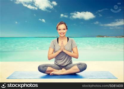fitness, sport, people and healthy lifestyle concept - woman making yoga meditation in lotus pose on mat over beach background. woman making yoga meditation in lotus pose on mat