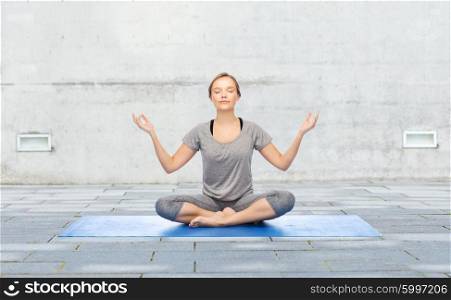 fitness, sport, people and healthy lifestyle concept - woman making yoga meditation in lotus pose on mat over urban street background