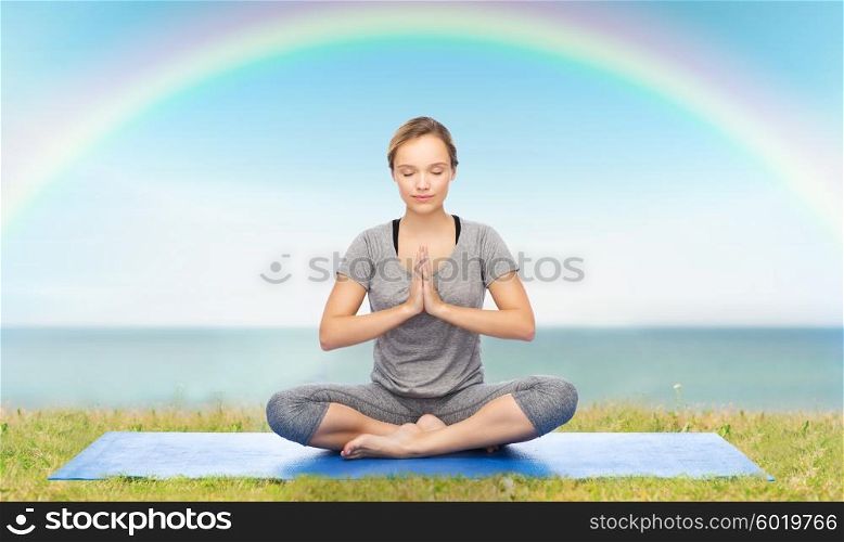 fitness, sport, people and healthy lifestyle concept - woman making yoga meditation in lotus pose on mat over blue sky, rainbow and sea background